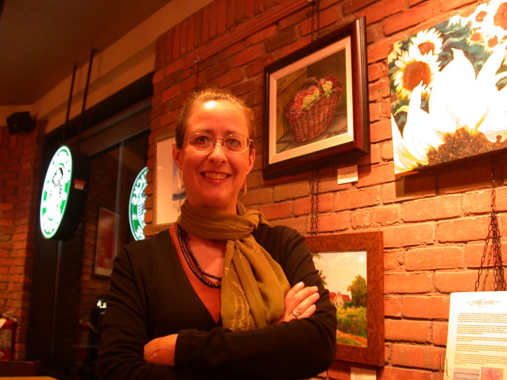 Image of Beth Himsworth next to art.  She has her arms folded.  She is wearing glasses, earrings, a brown scarf and black shirt with a black beaded necklace.